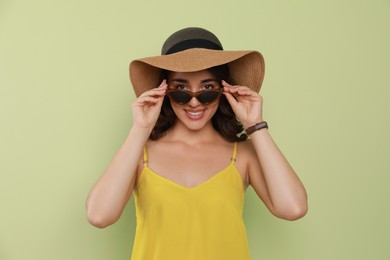 Photo of Beautiful young woman with straw hat and stylish sunglasses on light green background