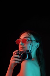 Beautiful woman with sunglasses and vintage camera posing in neon lights against black background