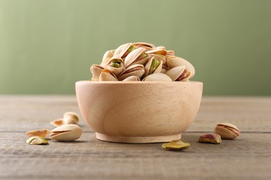 Photo of Tasty pistachios in bowl on wooden table against olive background, closeup