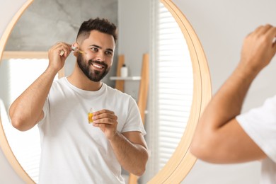 Photo of Handsome man applying cosmetic serum onto his face near mirror indoors
