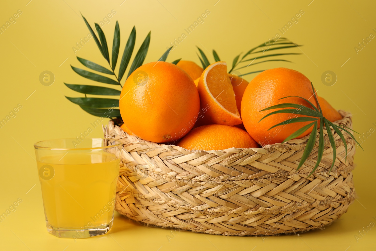 Photo of Fresh oranges in wicker basket and juice on yellow background, closeup