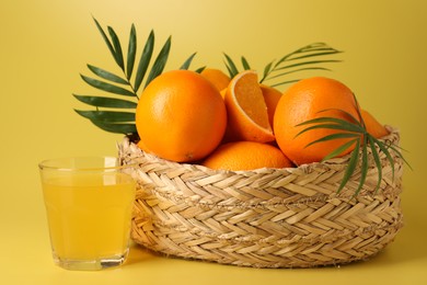 Fresh oranges in wicker basket and juice on yellow background, closeup