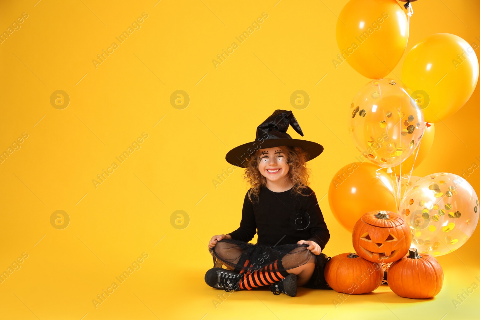 Photo of Cute little girl with pumpkins and balloons wearing Halloween costume on yellow background. Space for text