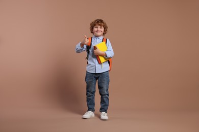 Photo of Happy schoolboy with backpack and books showing thumb up gesture on brown background