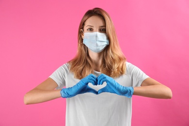 Young woman in medical gloves and protective mask making heart with hands on pink background
