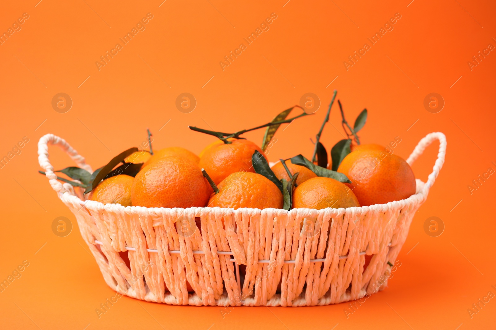 Photo of Fresh ripe tangerines and leaves in basket on orange table