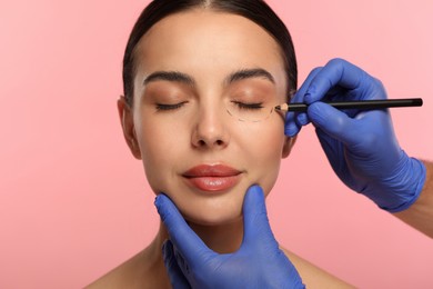 Doctor with pencil preparing patient for cosmetic surgery operation on pink background, closeup