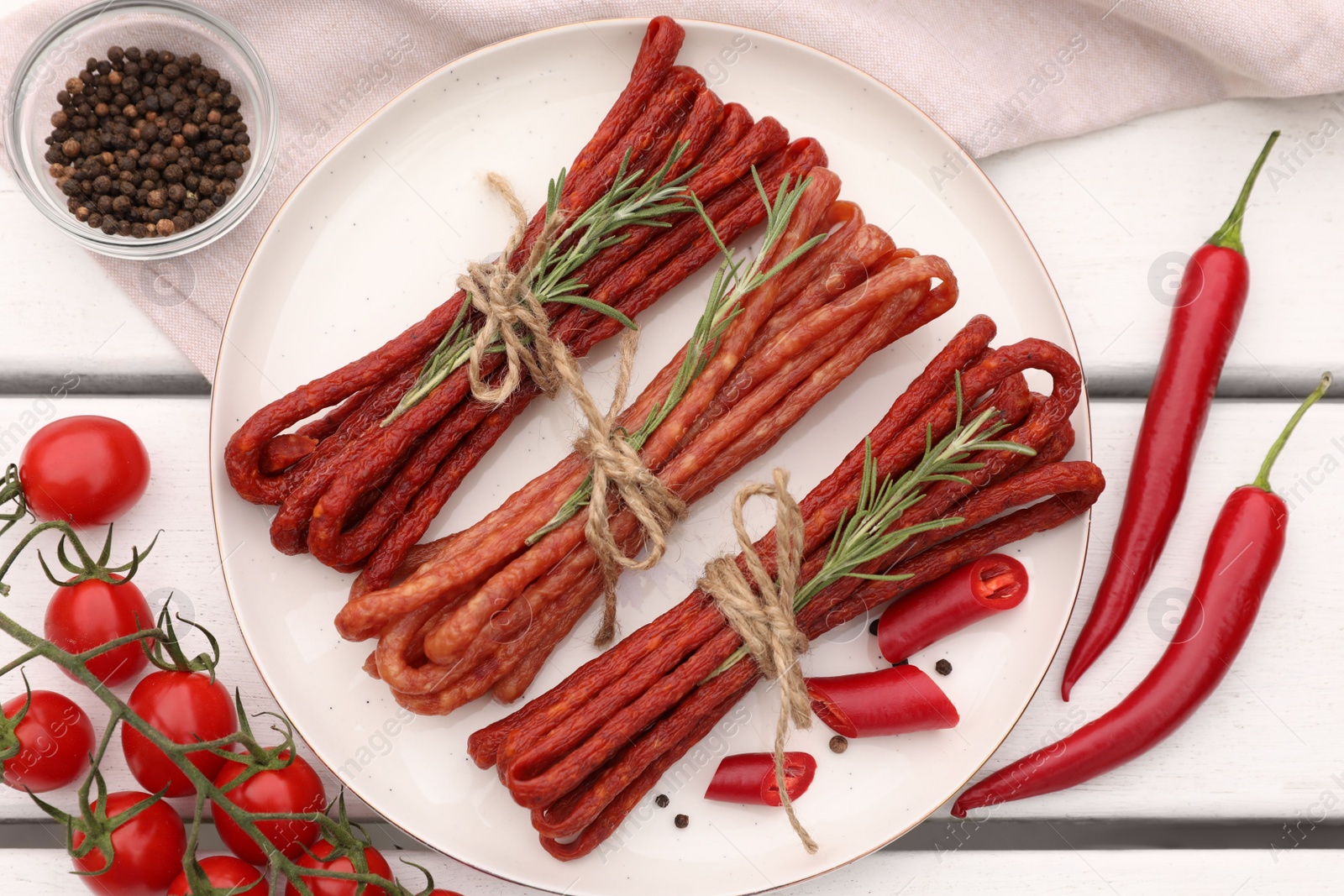 Photo of Bundles of delicious kabanosy with rosemary, peppercorn, chilli and tomatoes on white wooden table, flat lay