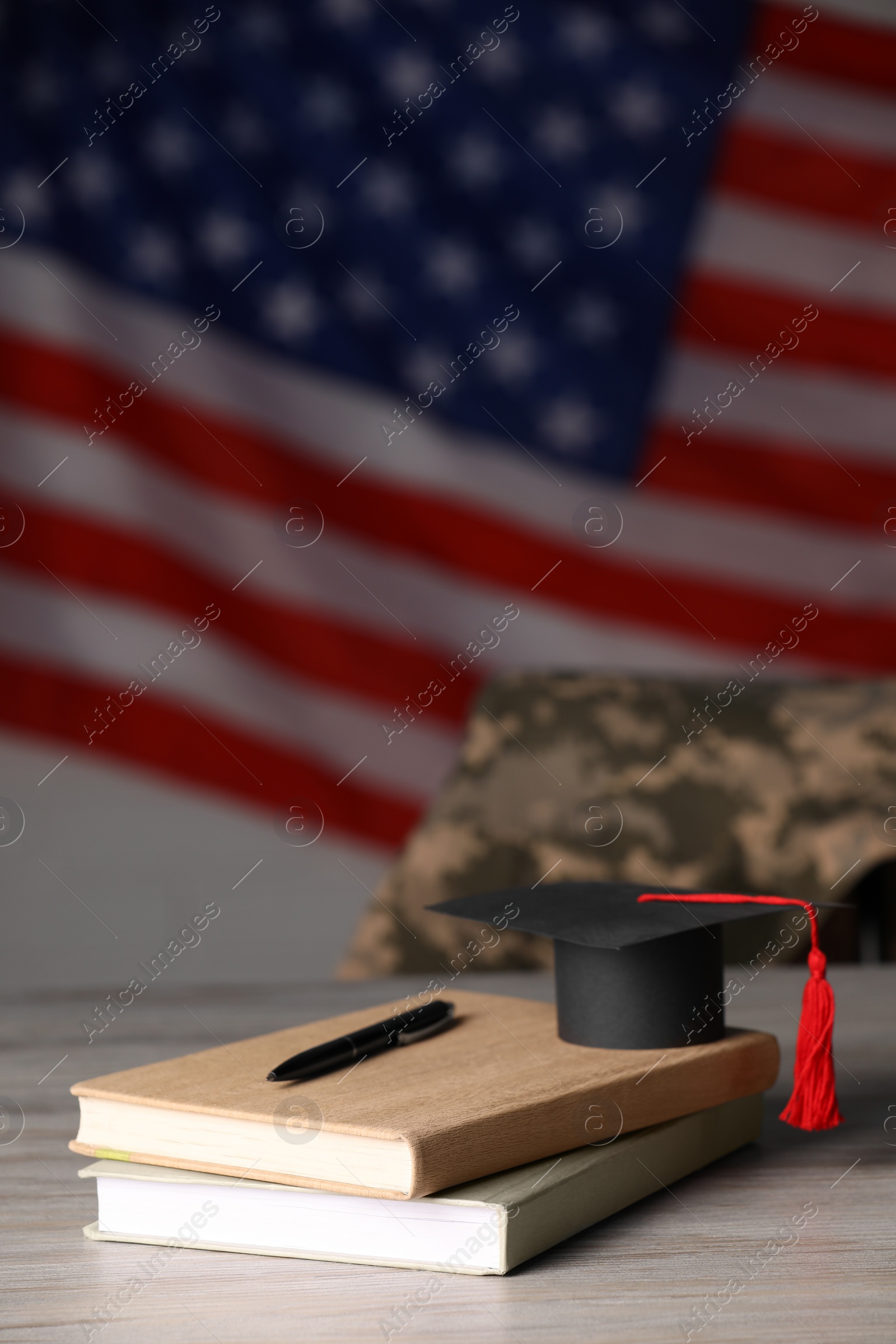 Photo of Notebooks, mortarboard and pen on wooden table against flag of USA. Military education