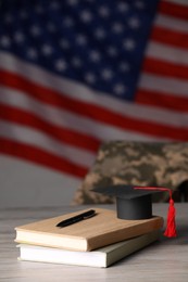 Photo of Notebooks, mortarboard and pen on wooden table against flag of USA. Military education