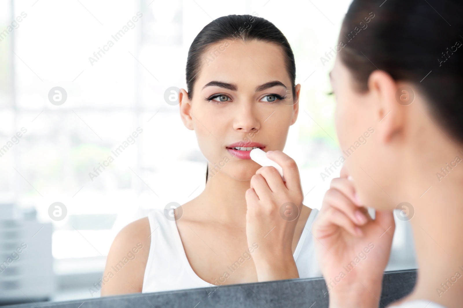 Photo of Young woman applying balm on her lips near mirror indoors