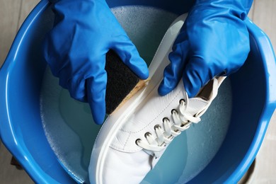 Woman with gloves and sponge cleaning stylish sneakers in wash basin, top view