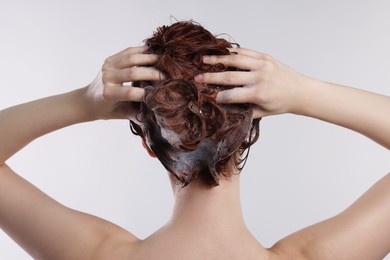 Young woman washing her hair with shampoo on light grey background, back view