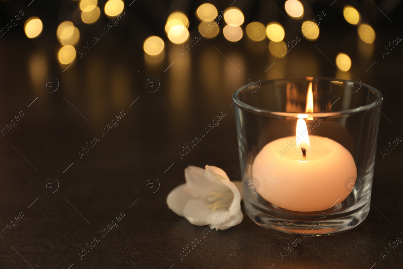 Photo of Burning candle in glass holder and white flower on table against blurred lights, space for text