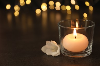 Photo of Burning candle in glass holder and white flower on table against blurred lights, space for text