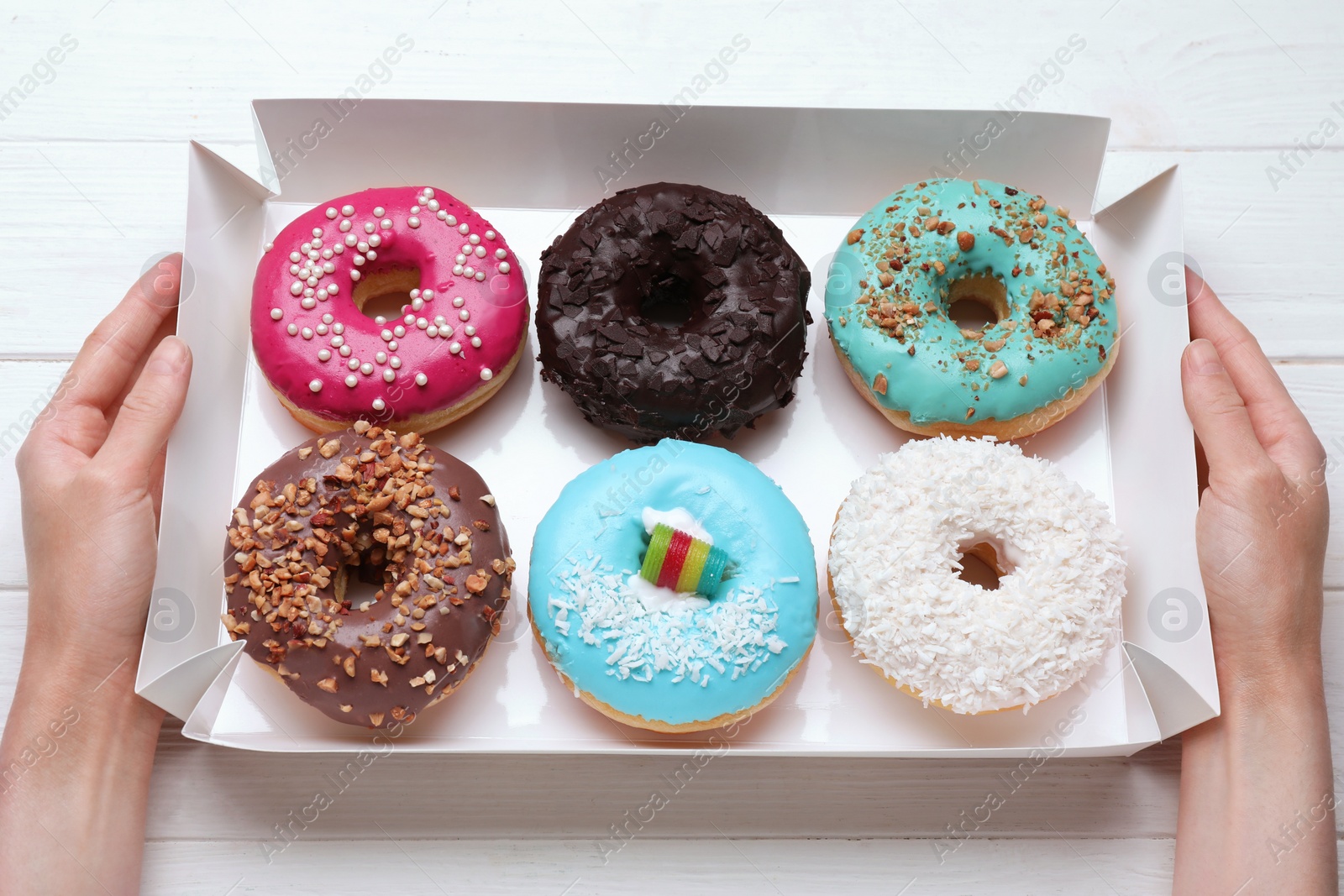 Photo of Woman holding box with tasty glazed donuts at white wooden table, top view