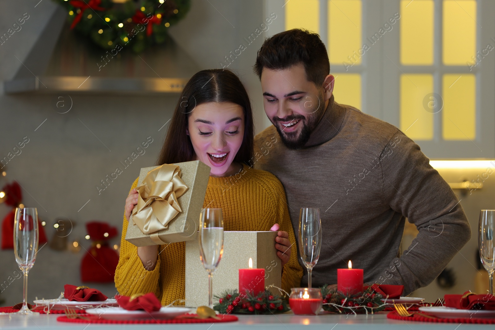 Photo of Surprised young woman opening Christmas gift from her boyfriend at table in kitchen