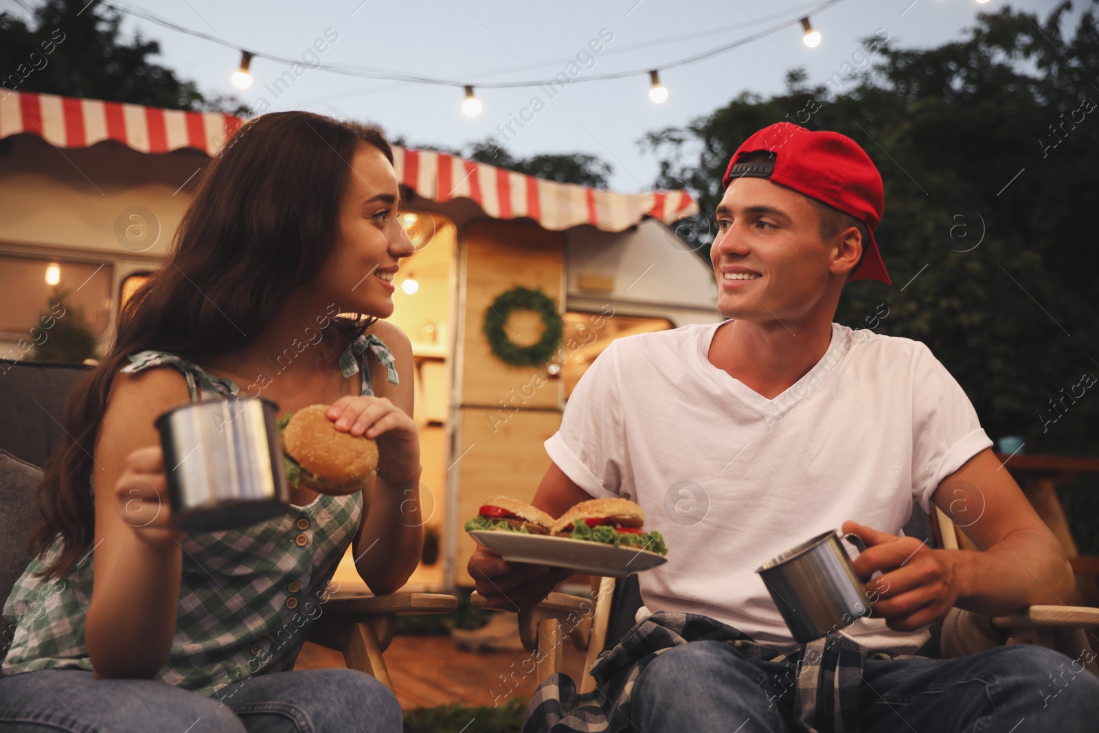 Photo of Happy couple with burgers and cups resting near motorhome. Camping season