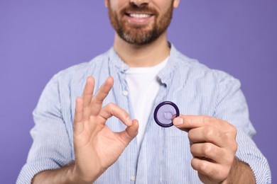 Photo of Man with condom showing ok gesture on purple background, closeup. Safe sex