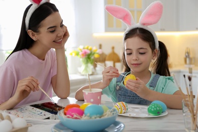 Happy mother with her cute daughter painting Easter eggs at table in kitchen