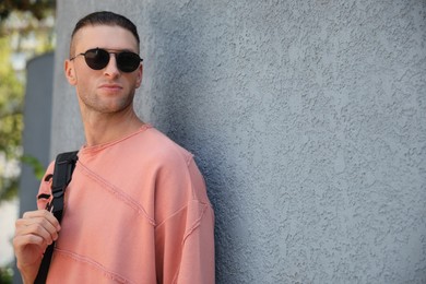 Photo of Handsome young man in stylish sunglasses with backpack near grey wall outdoors, space for text