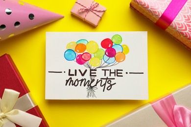 Photo of Life-affirming phrase Live The Moments. Flat lay composition with card and gift boxes on yellow background