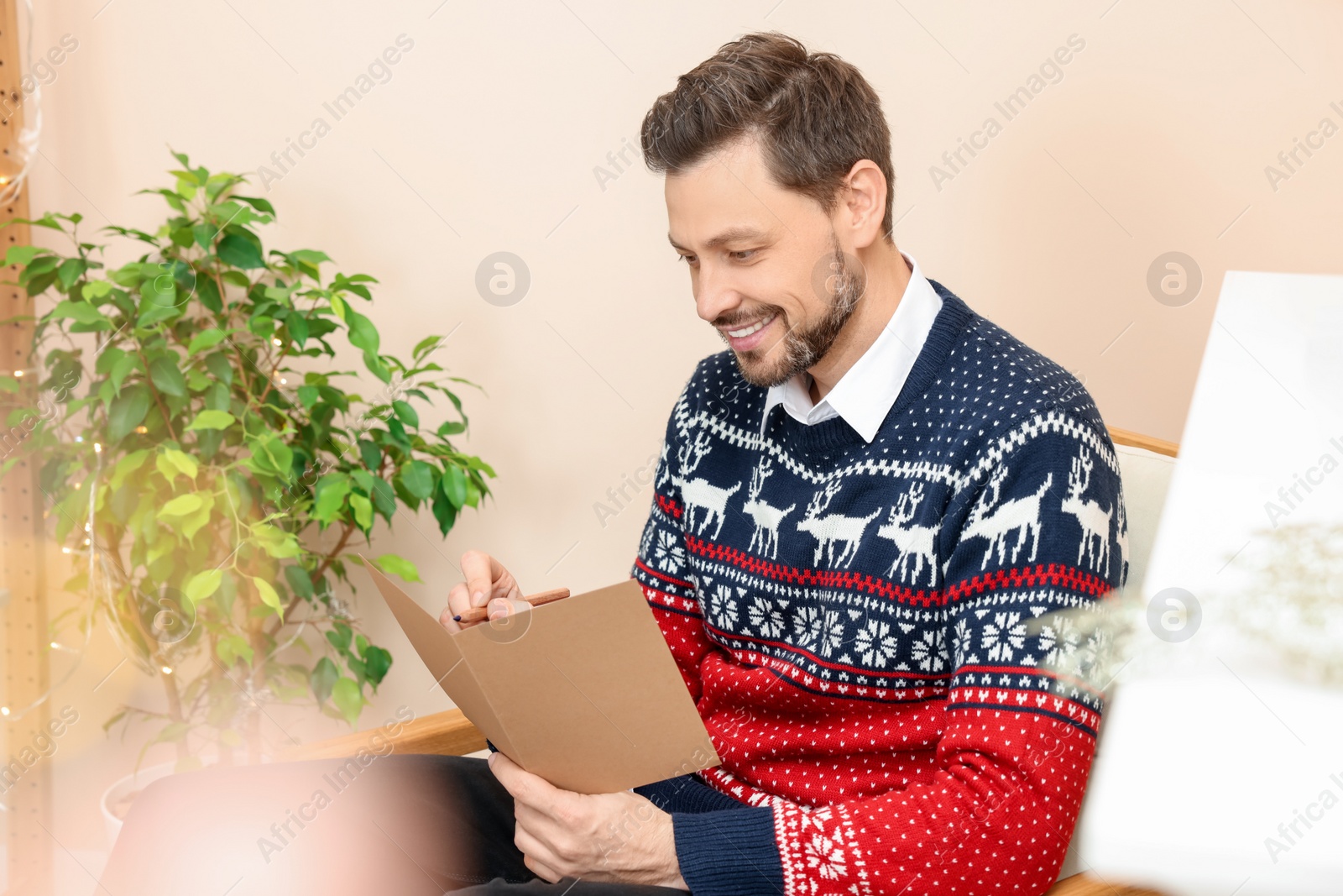 Photo of Happy man writing wishes in Christmas greeting card in living room