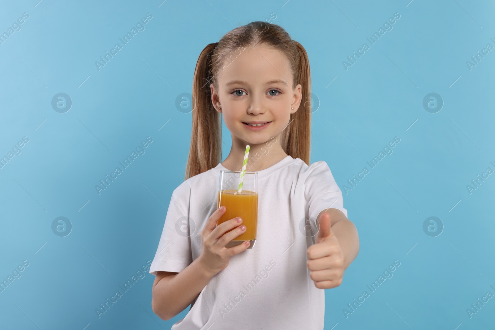 Photo of Cute little girl with fresh juice showing thumbs up on light blue background
