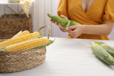 Photo of Woman husking corn cob at white wooden table, focus on basket