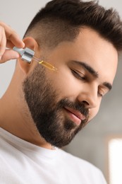 Handsome man applying cosmetic serum onto his face on blurred background, closeup