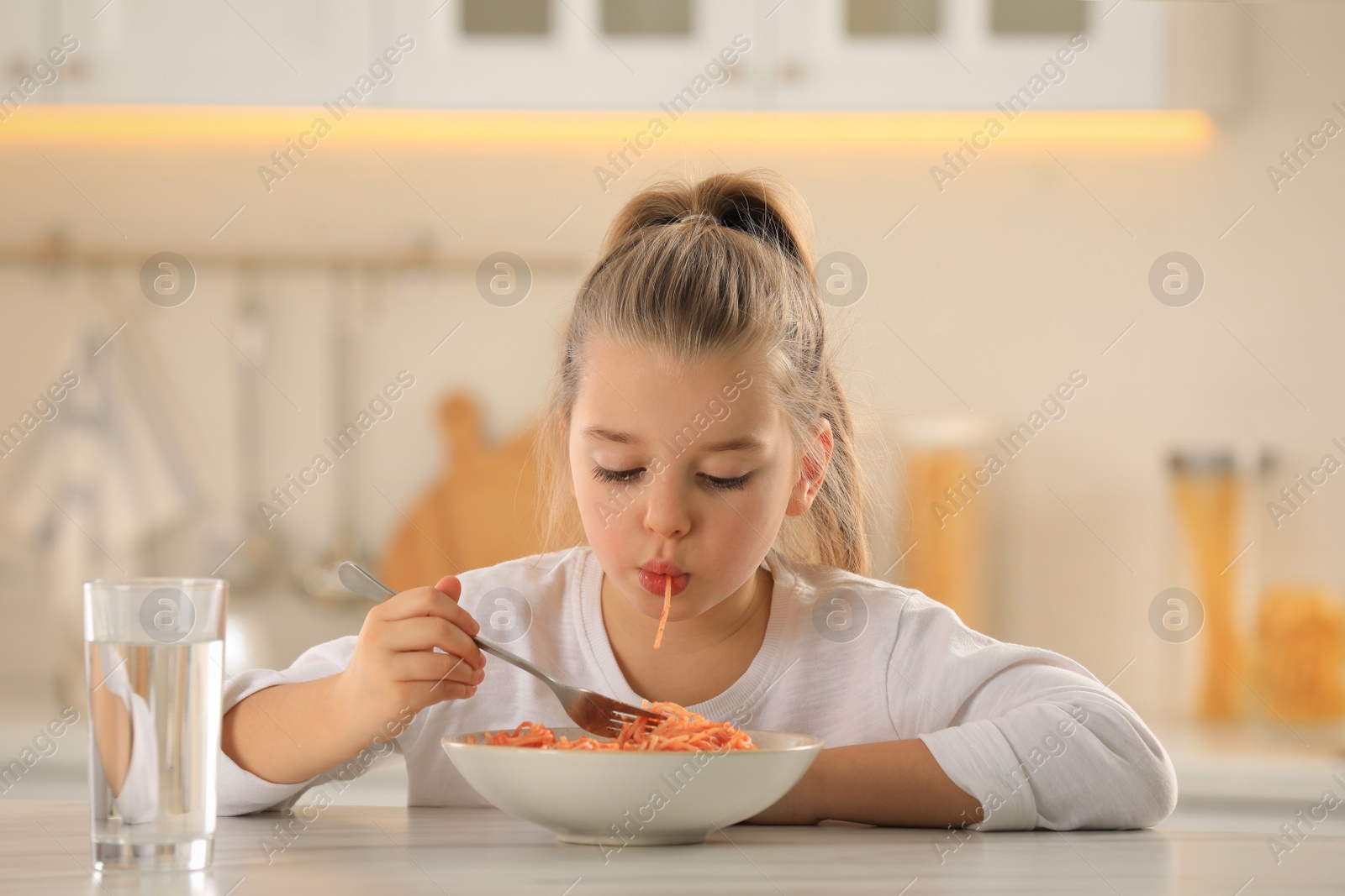 Photo of Cute little girl eating tasty pasta at table in kitchen