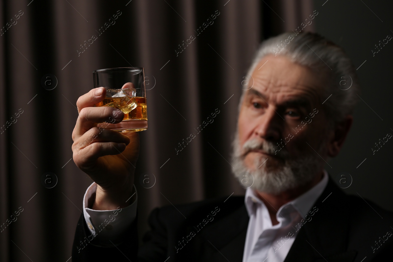 Photo of Senior man in suit holding glass of whiskey with ice cubes on brown background, selective focus