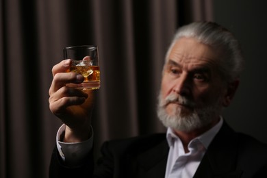 Photo of Senior man in suit holding glass of whiskey with ice cubes on brown background, selective focus