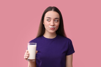 Cute woman with milk mustache holding glass of tasty dairy drink on pink background