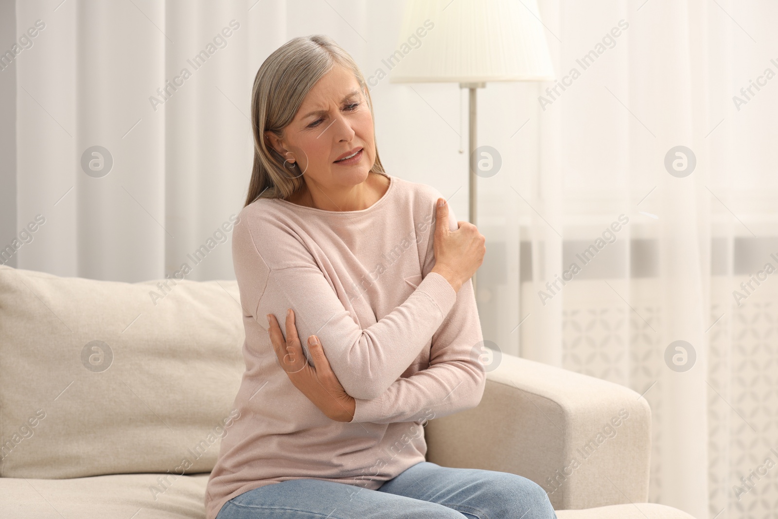 Photo of Mature woman suffering from pain in arm on sofa at home. Rheumatism symptom