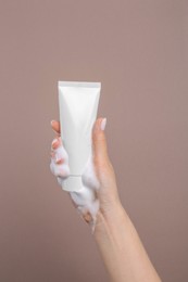 Photo of Woman with cleansing foam on hand holding tube of cosmetic product against brown background, closeup