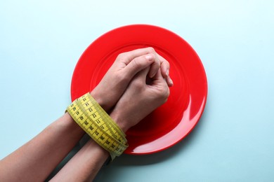 Woman tied with measuring tape above plate against light blue background, top view. Diet concept