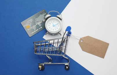 Shopping cart with tag, credit card and alarm clock on color background, flat lay