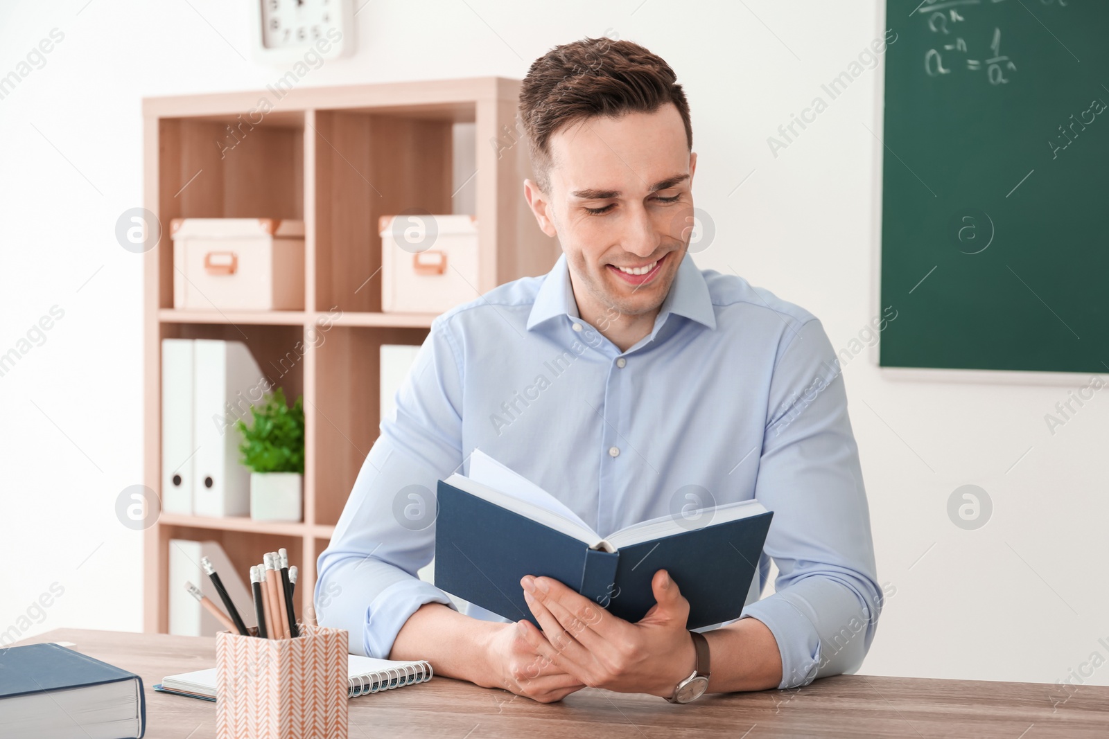 Photo of Young male teacher with notebook sitting at table in classroom