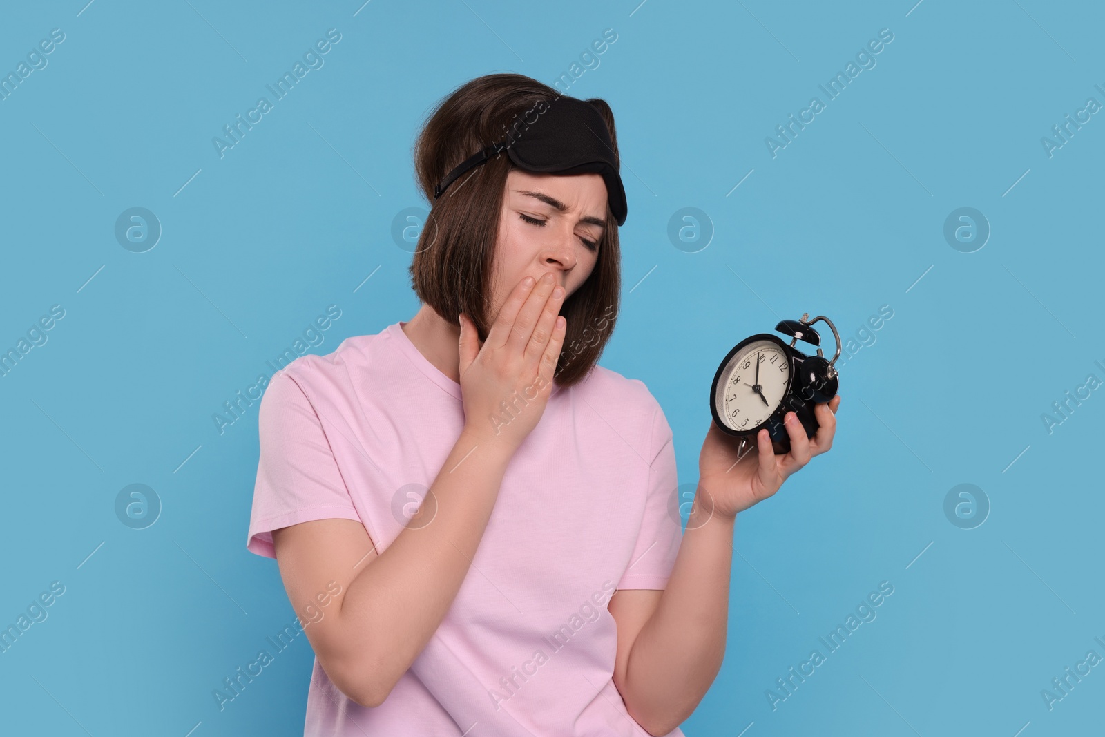 Photo of Sleepy young woman with sleep mask and alarm clock yawning on light blue background. Insomnia problem