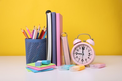 Photo of Different school stationery and alarm clock on white table against yellow background. Back to school
