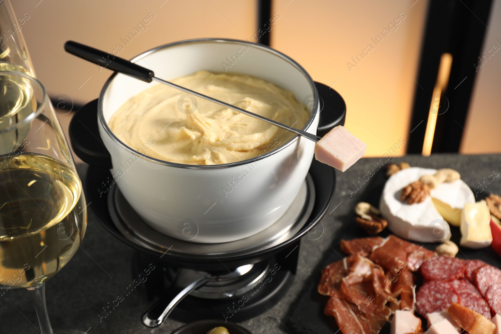 Photo of Fork with piece of ham, fondue pot with melted cheese, wine and snacks on grey table, closeup