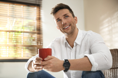 Young man with cup of drink relaxing near window at home