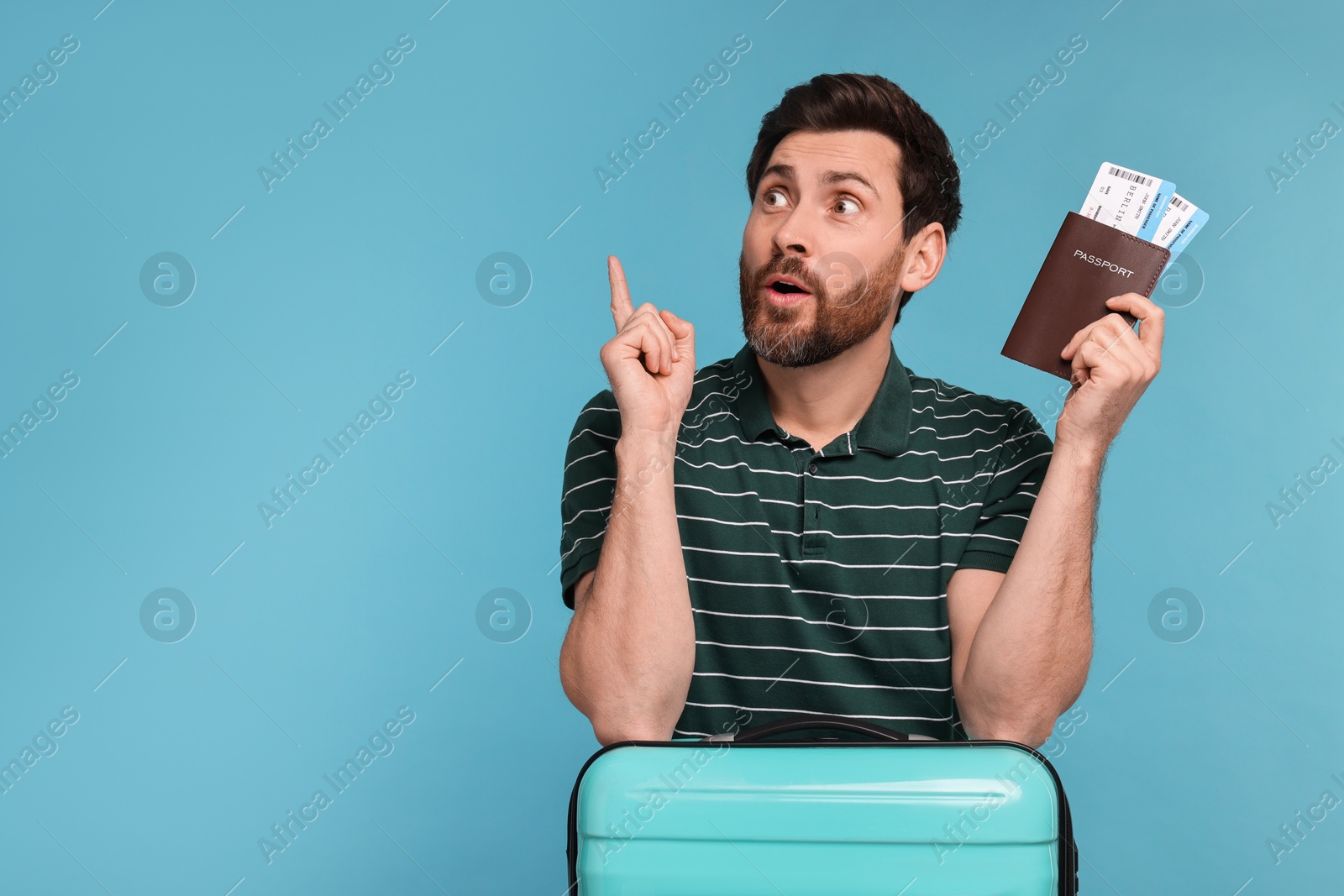 Photo of Surprised man with passport, suitcase and tickets pointing at something on light blue background. Space for text