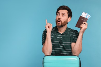 Surprised man with passport, suitcase and tickets pointing at something on light blue background. Space for text