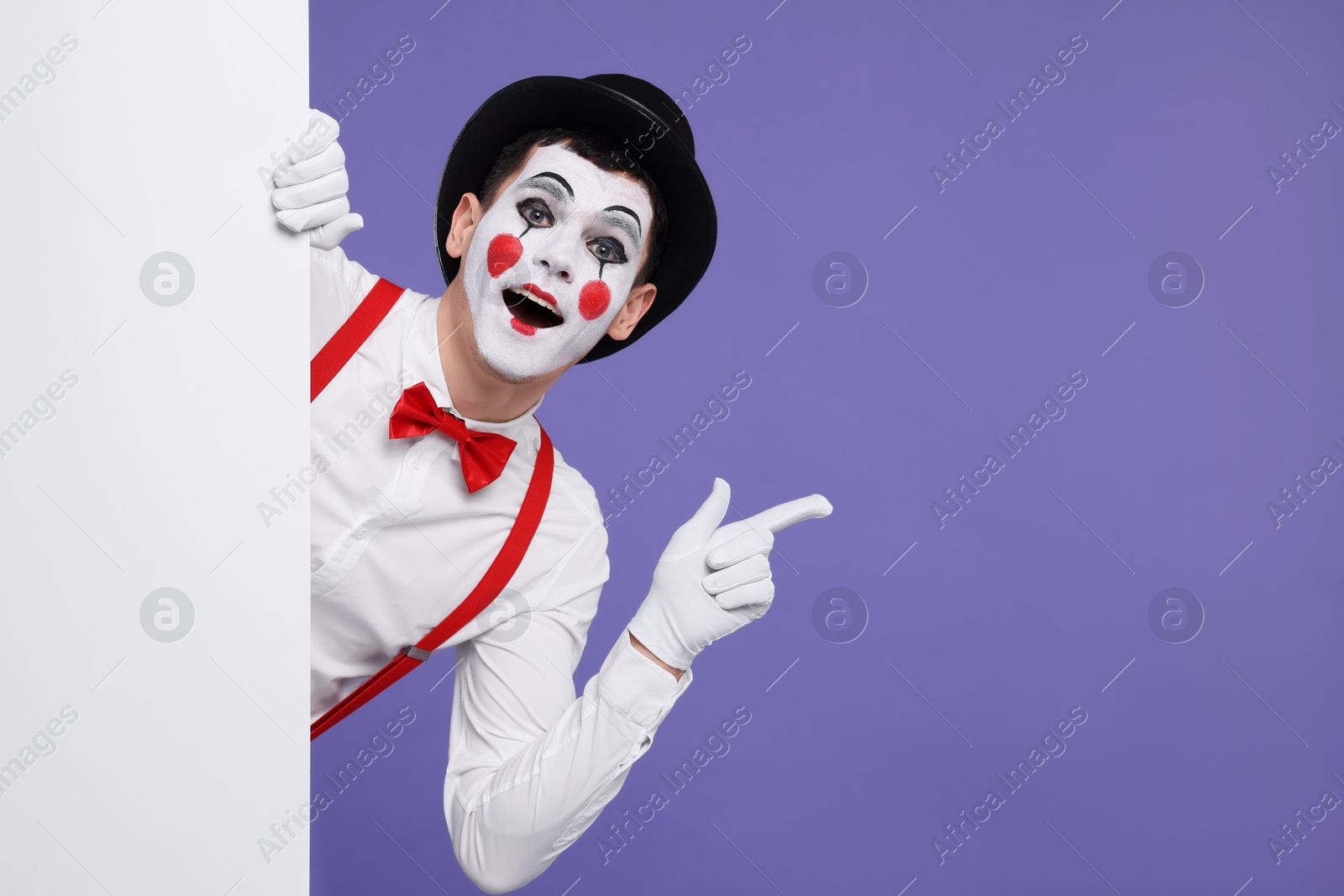 Photo of Funny mime artist peeking out of blank poster and pointing at something on purple background. Space for text
