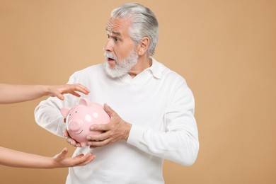 Photo of Woman taking piggy bank from confused senior man on beige background. Be careful - fraud