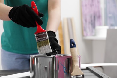 Woman dipping brush into can of white paint at table indoors, closeup. Space for text
