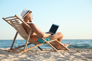 Young man with laptop in deck chair on beach