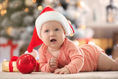 Photo of Little baby in Santa hat with Christmas decoration on floor at home
